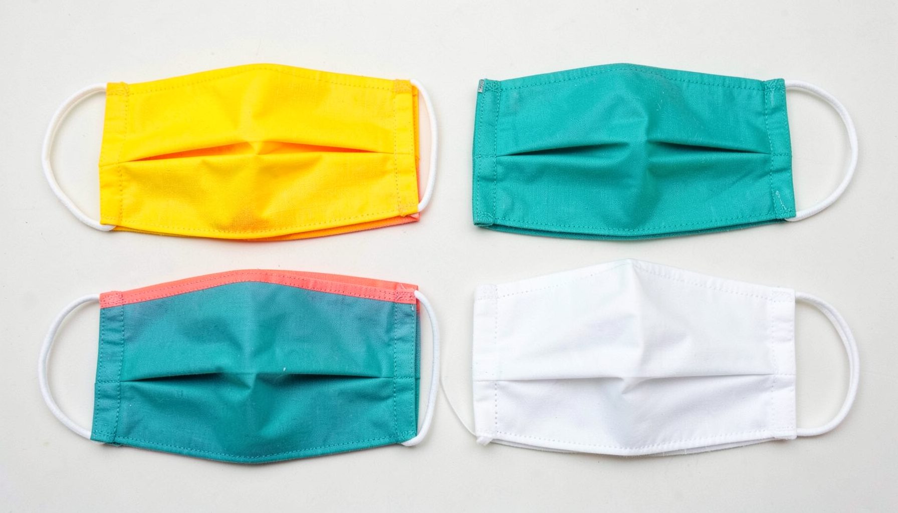 Quatro máscaras faciais de tecido em tons amarelo, azul-petróleo e branco dispostas em grelha sobre fundo claro.