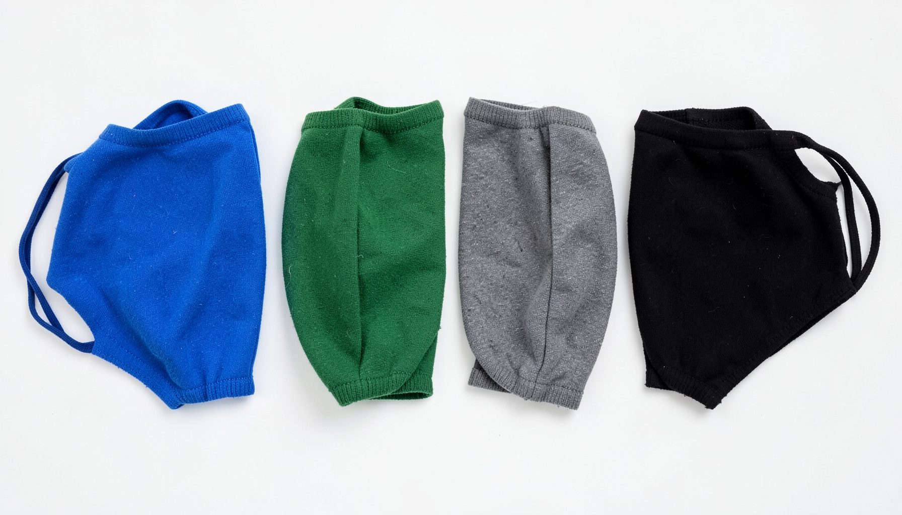 Quatro máscaras faciais de malha dobradas nas cores azul, verde, cinza e preto alinhadas sobre fundo branco, disposição limpa e ordenada.