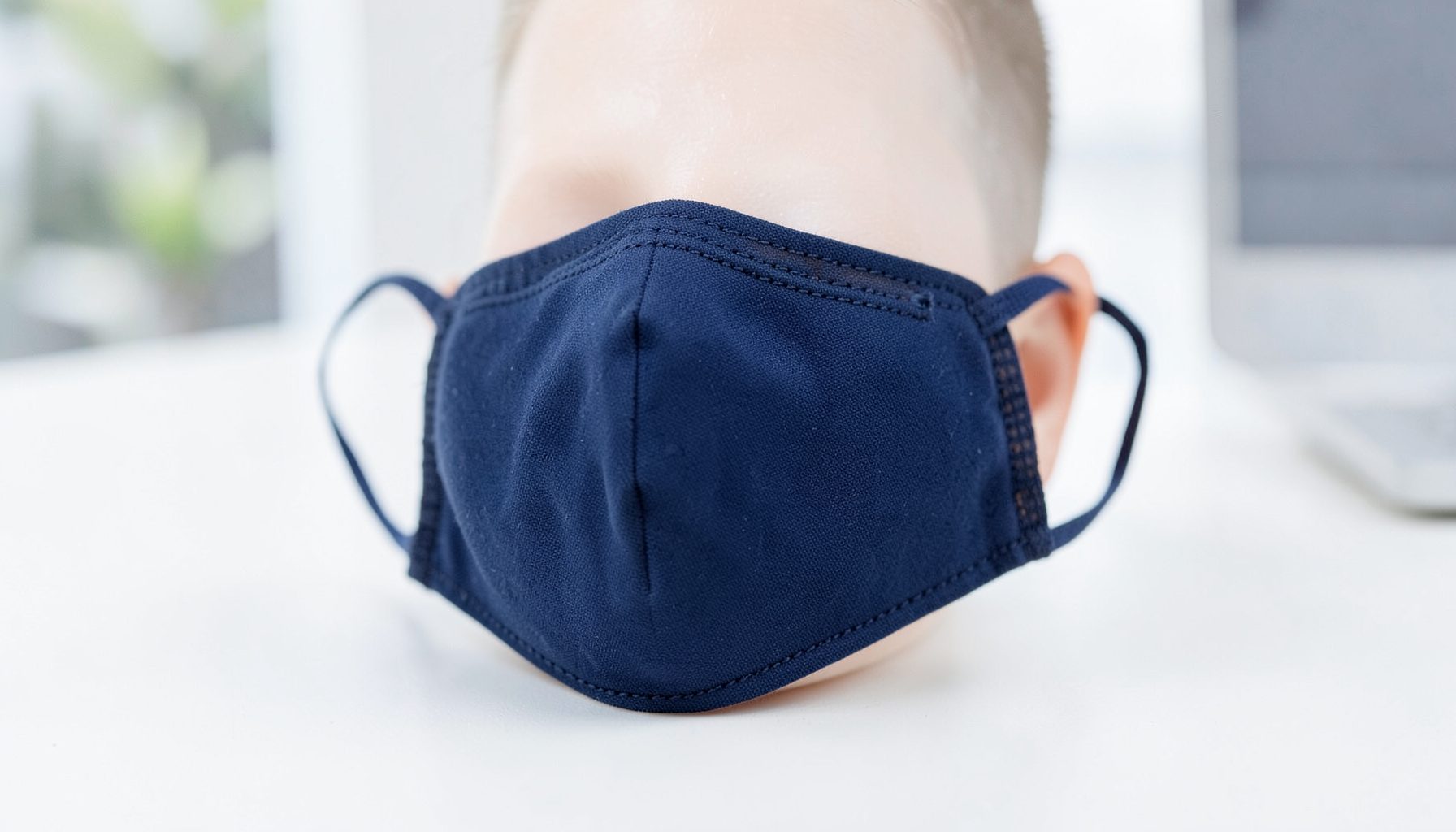 Máscara facial têxtil em tom azul-escuro sobre superfície clara, com costura visível e elásticos de orelha, ambiente limpo e corporativo.