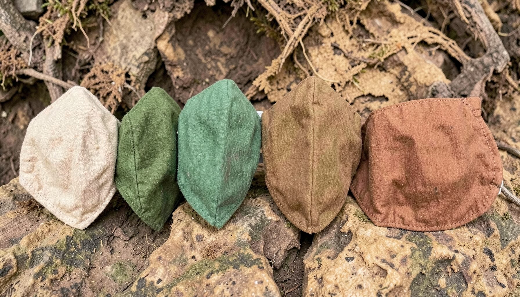 Máscaras faciais de tecido em tons creme, verde e ferrugem dispostas sobre uma superfície natural de raízes e pedra, com atmosfera terrosa e artesanal.