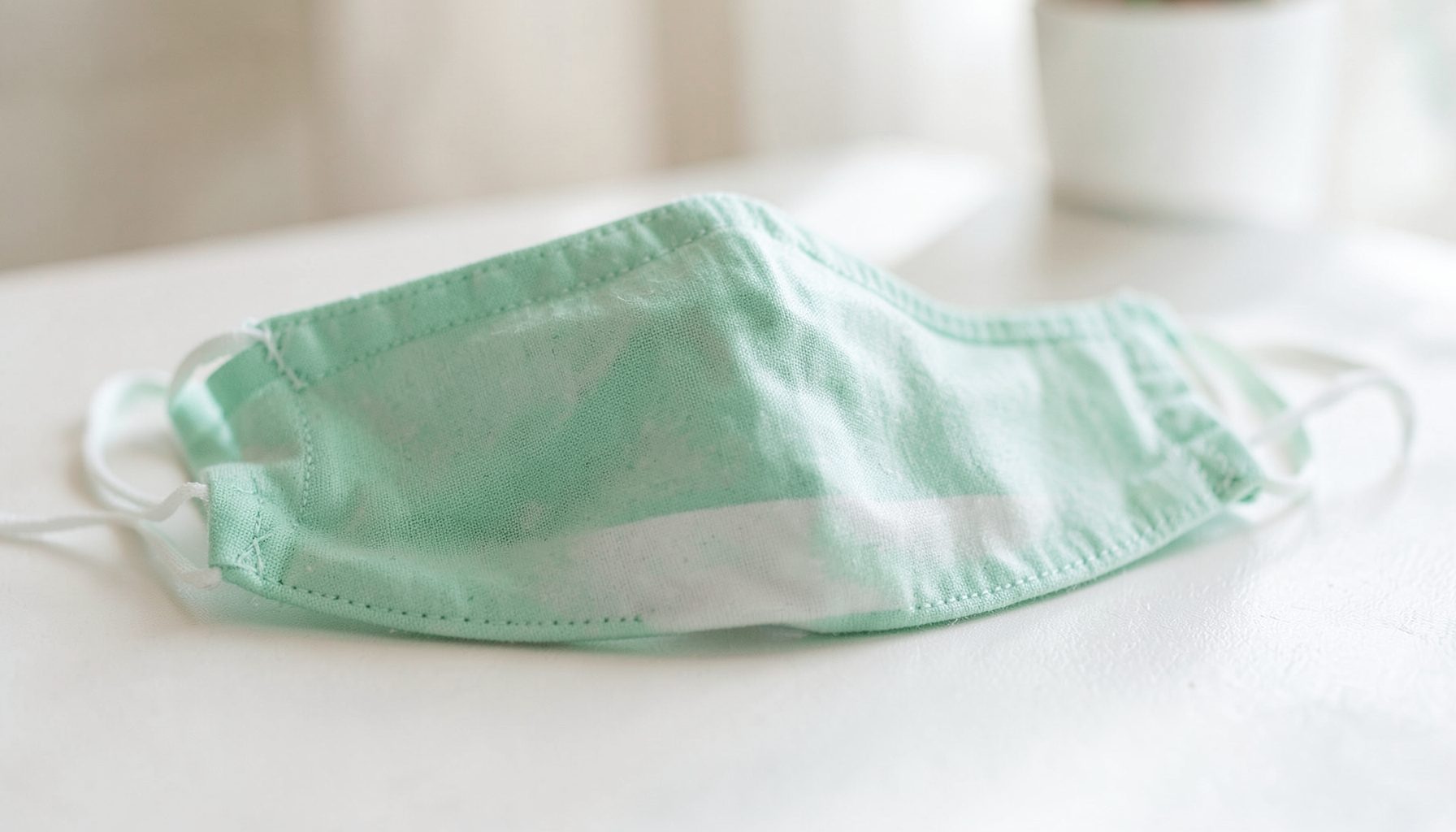 Máscara facial em tecido verde menta com padrão branco e cordões finos, sobre mesa clara