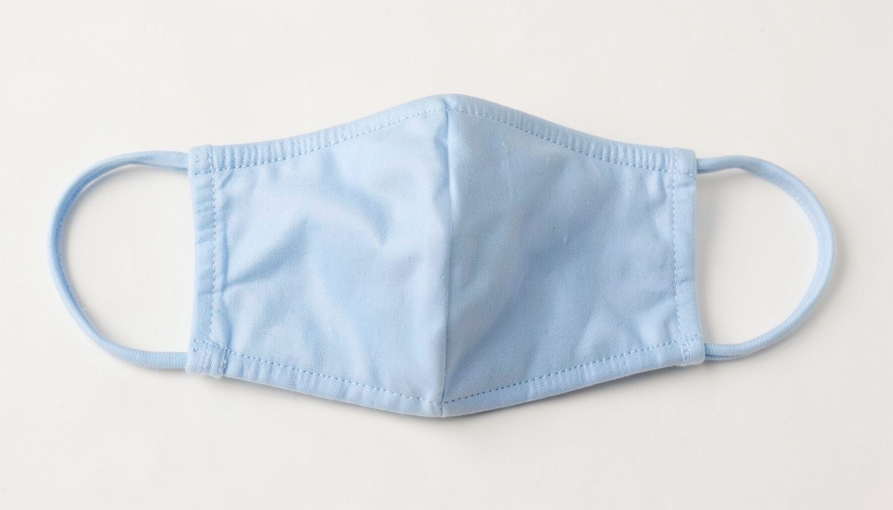 Máscara facial de tecido azul-claro dobrada com elásticos para as orelhas sobre fundo branco.