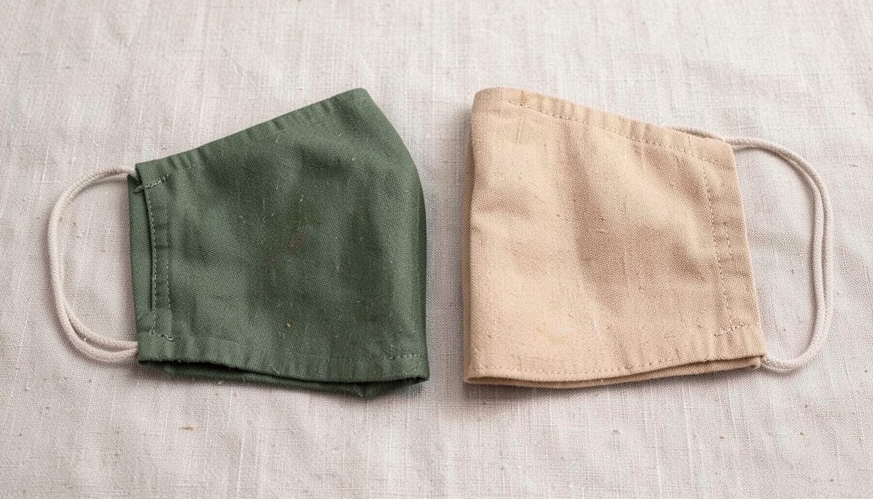 Duas máscaras faciais de tecido dobradas, uma verde-azeitona e outra bege, sobre uma superfície clara texturizada, tom minimalista e natural.